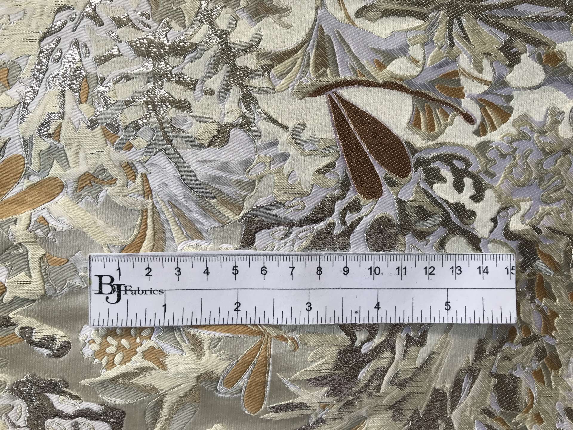 ruler sits on top of metallic brocade with florals in ivory light gold and silver to show scale of pattern