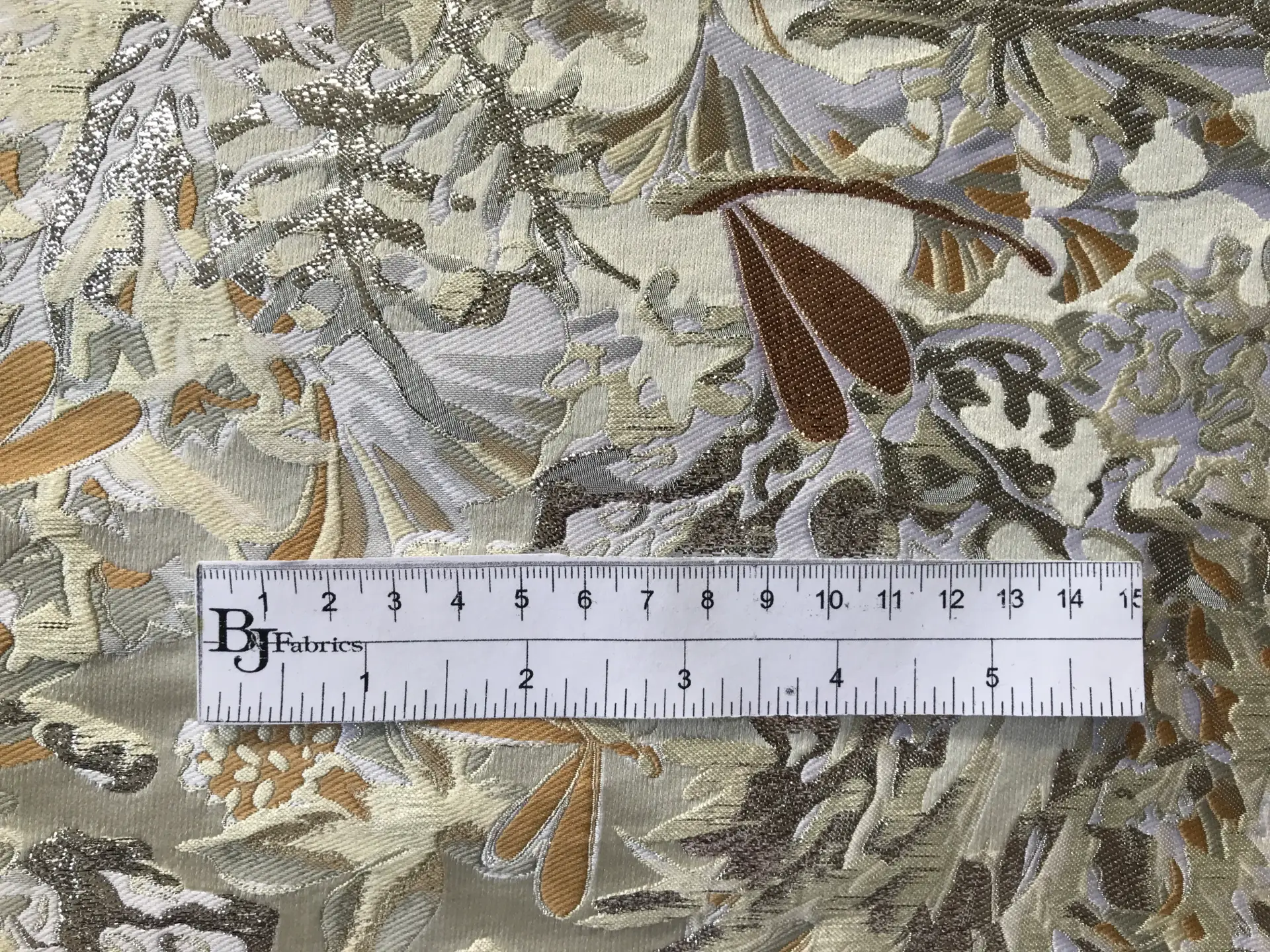 ruler sits on top of metallic brocade with florals in ivory light gold and silver to show scale of pattern
